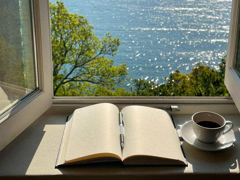 An open notebook and a cup of coffee on a sunny window seat over the lake